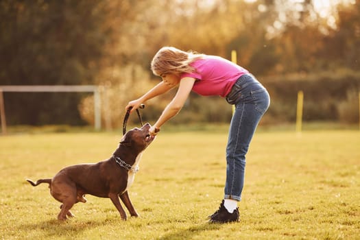 Playing together. Woman in casual clothes is with pit bull outdoors.