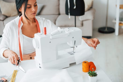 Working by using sewing machine. Seamstress is in her office that is with different clothes.