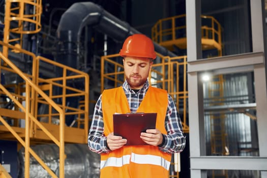 With notepad. Construction worker in uniform is in the factory.