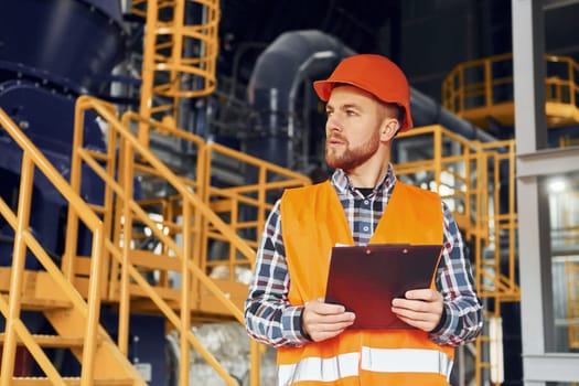 With notepad. Construction worker in uniform is in the factory.