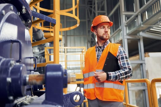 With notepad. Construction worker in uniform is in the factory.
