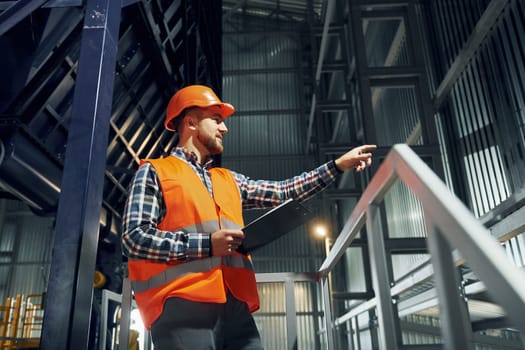In orange colored hard hat. Construction worker in uniform is in the factory.
