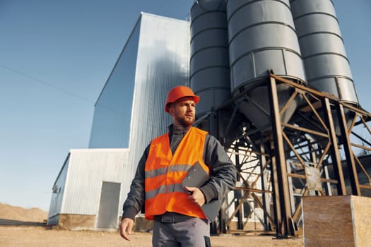 Holding notepad in hands. Construction worker in uniform is outdoors near the factory.