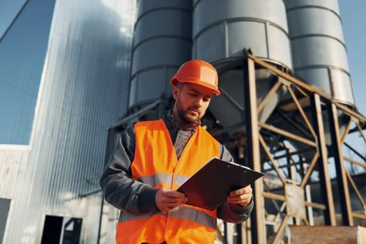 Holding notepad in hands. Construction worker in uniform is outdoors near the factory.