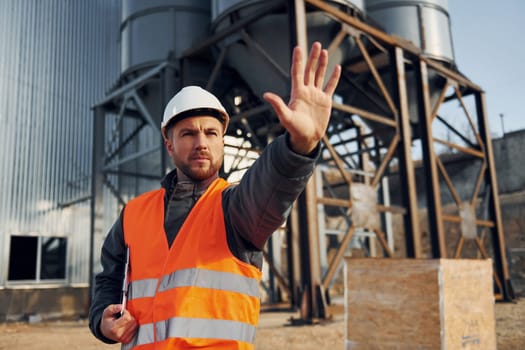 Showing stop gesture. Construction worker in uniform is outdoors near the factory.