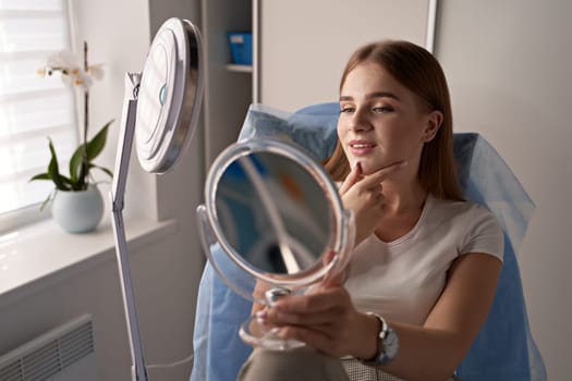 Young beautiful female customer with mirror getting ready for beauty procedures in a medical salon