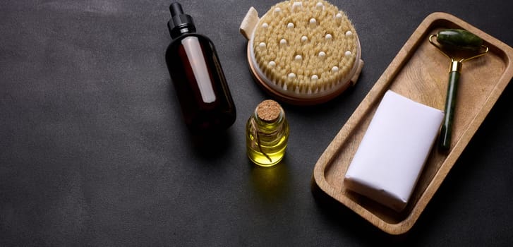 Brown glass bottle for cosmetics, a piece of soap wrapped in paper and a brush for dry body massage on a black background