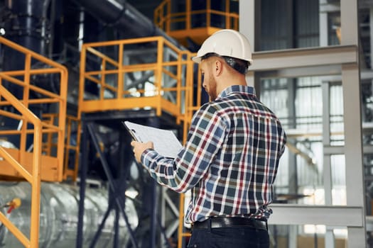 In white hard hat. Construction worker in uniform is in the factory.