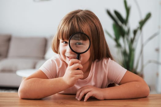 Having fun. Little girl is sitting by the table with magnifying glass.