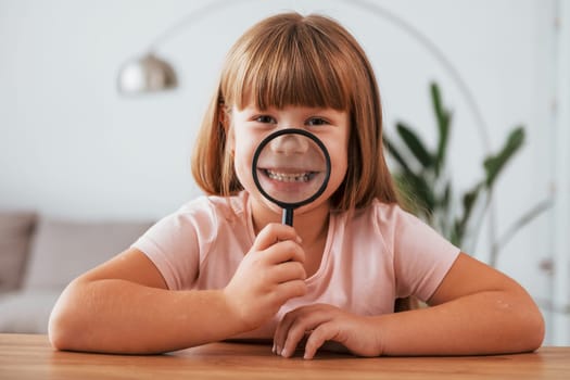 Having fun. Little girl is sitting by the table with magnifying glass.