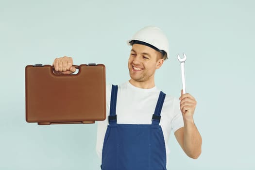 Holding brown case. Worker in uniform and hard hat standing in the studio with equipment.