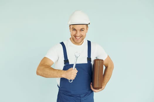 Holding brown case. Worker in uniform and hard hat standing in the studio with equipment.