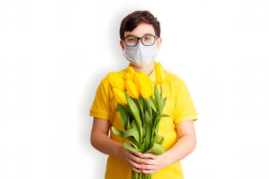 a brunette boy , dressed in a yellow T-shirt, holds a bouquet of yellow tulips ,in medical mask on his face, look at camera, on a white background. To close up.