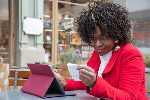 a young african american woman in a red coat sits at a table in a cafe and checking bill after shopping using a tablet, shocked by big expenses sitting with her mouth open, financial problems crisis