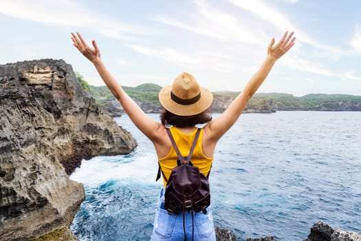 Back view of happy woman with arms up enjoying vacation on Nusa Penida, Bali. Female enjoying freedom in nature. Trip, vacation, mental health and healthy lifestyle concept.