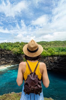 Rear view of young woman traveler standing on cliff looking at Broken Beach Nusa Penida Bali. Summer vacation. Traveling, freedom and wellness concept. Vertical image.