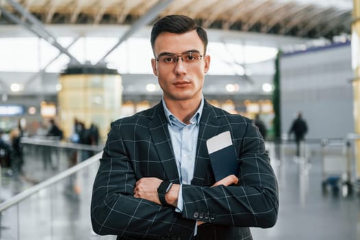 With arms crossed. Young businessman in formal clothes is in the airport at daytime.