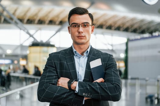 With arms crossed. Young businessman in formal clothes is in the airport at daytime.