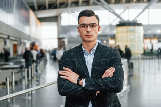 With arms crossed. Young businessman in formal clothes is in the airport at daytime.