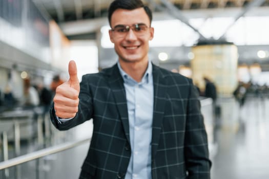 Showing thumb up. Young businessman in formal clothes is in the airport at daytime.