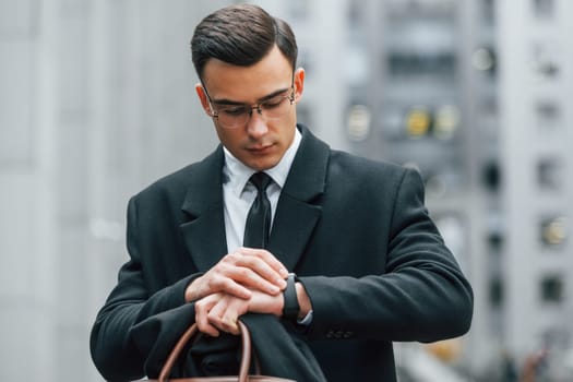 Looking at clock. Businessman in black suit and tie is outdoors in the city.