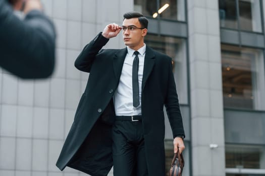 Posing for photographer. Businessman in black suit and tie is outdoors in the city.