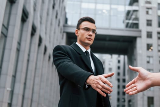 Doing handshake. Businessman in black suit and tie is outdoors in the city.