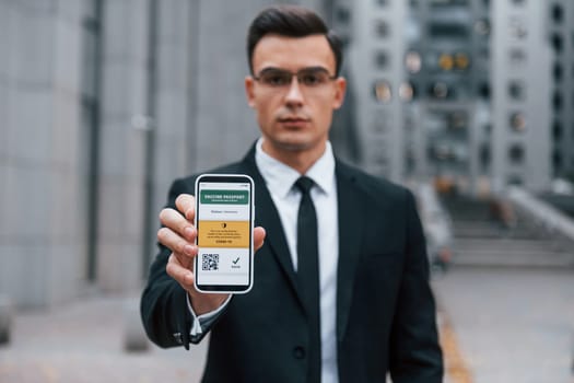 Holding phone with vaccination certificate. Businessman in black suit and tie is outdoors in the city.