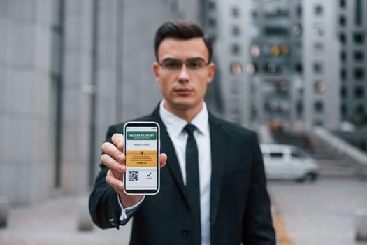 Holding phone with vaccination certificate. Businessman in black suit and tie is outdoors in the city.
