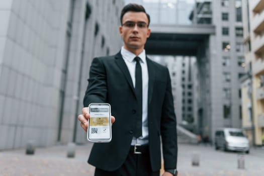 Holding phone with vaccination certificate. Businessman in black suit and tie is outdoors in the city.