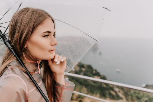 Woman rain park. Happy woman portrait wearing a raincoat with transparent umbrella outdoors on rainy day in park near sea. Girl on the nature on rainy overcast day