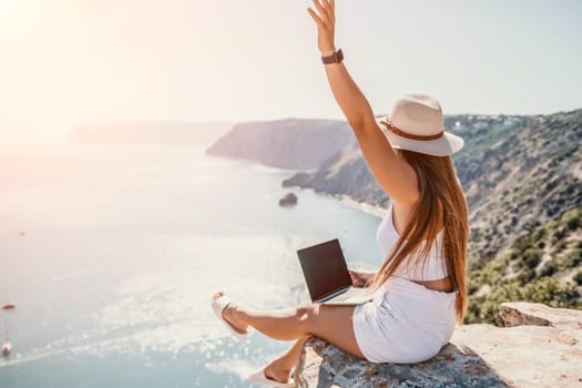 Successful business woman in yellow hat working on laptop by the sea. Pretty lady typing on computer at summer day outdoors. Freelance, travel and holidays concept.