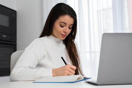 Intelligent female student writing notes to textbook sitting at home and preparing to the exam.