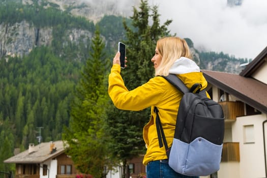 Blonde tourist takes photos using smartphone of misty Alpine mountains. Woman makes photos of mountain hills covered with clouds