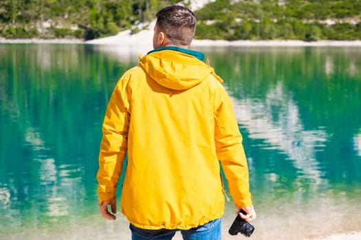 Tourist in yellow sport jacket holds a camera and admires the scenery of Lake Braies and mountains in Italy, back view.