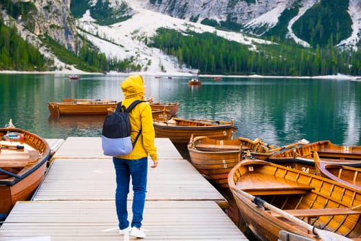 Inspiring female traveler in yellow jacket visits popular lake Braies and stands on the pier with wooden boats and the cinematic mountains of Dolomites Alps in autumn