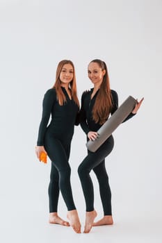 Holding mat. Two young women in sportive clothes is together indoors against white background.