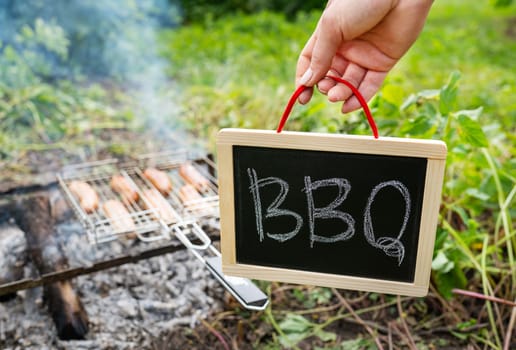 Close-up of fried sausages on the grill. A man is holding a sign with the inscription barbecue. Rest in nature, picnic, weekend
