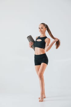 Holding yoga mat and standing. Young woman in sportive clothes doing gymnastics indoors.