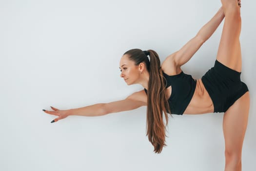 Standing against white background. Young woman in sportive clothes doing gymnastics indoors.