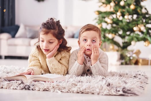 Laying down on the ground. Little brother and sister is at christmas decorated room together.