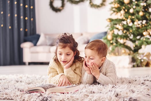 Laying down on the ground. Little brother and sister is at christmas decorated room together.