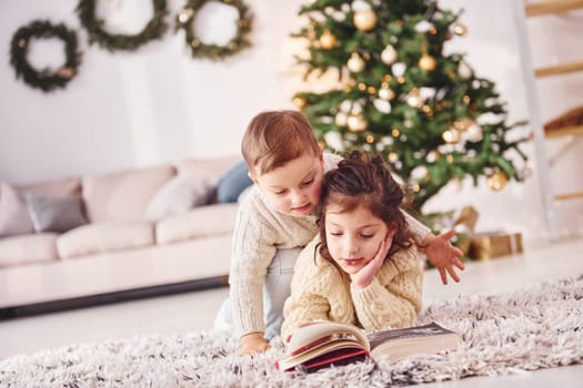 Laying down on the ground. Little brother and sister is at christmas decorated room together.