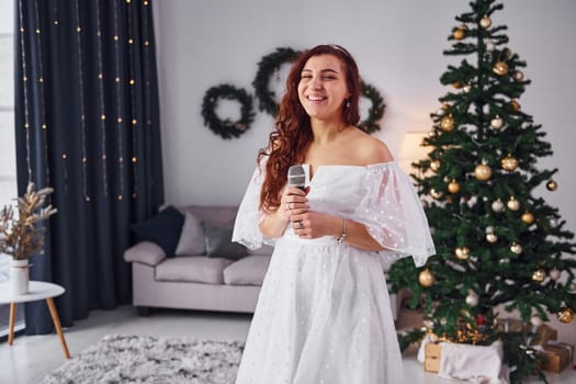 Woman in white dress and with microphone in hands is singing in the christmas decorated domestic room.