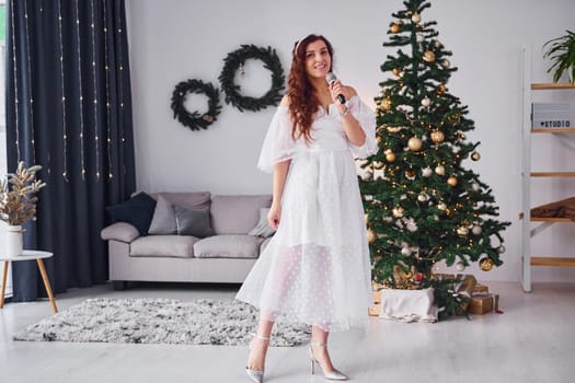 Woman in white dress and with microphone in hands is singing in the christmas decorated domestic room.