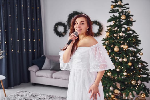 Standing in the christmas decorated domestic room. Woman in white dress and with microphone in hands is singing in the .
