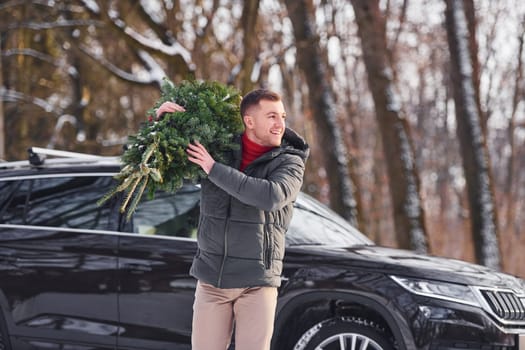 Front view. Man with little green fir is outdoors near his car. Conception of holidays.