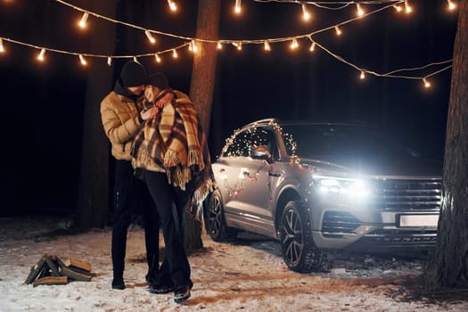 Standing near car. Couple is in the forest and celebrating New year.