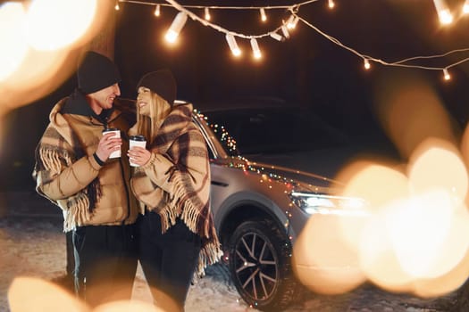 Standing near car. Couple is in the forest and celebrating New year.