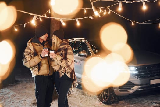 Standing near car. Couple is in the forest and celebrating New year.